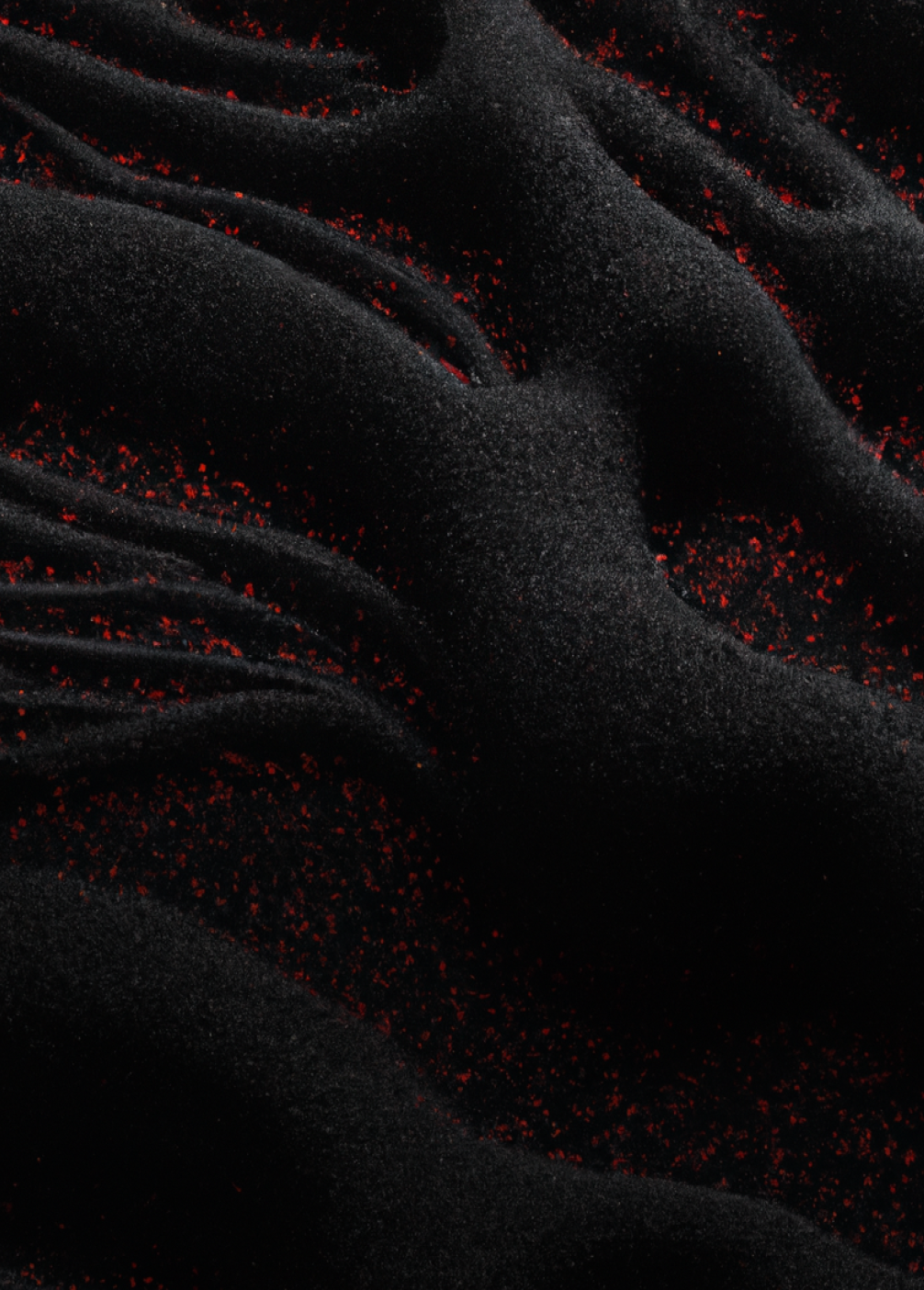 A textured black surface with flowing, organic ridges and scattered red specks, resembling sand dunes or fabric folds.